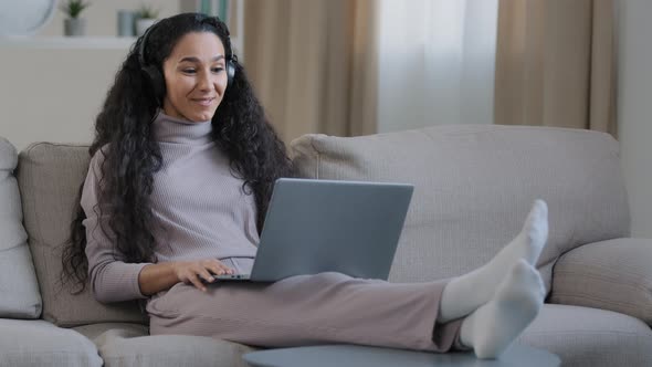 Hispanic Arabian Cute Woman Girl Wear Headphones Sit on Sofa at Home Living Room Hold Laptop Use