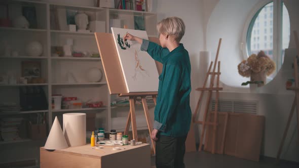 A Young Blonde Woman Artist Painting Upper Branches of Tree in Green Color in the Art Studio