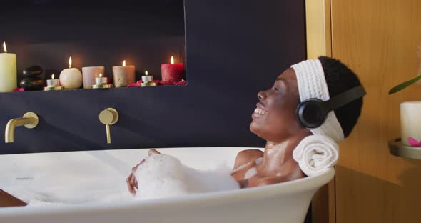 Smiling african american woman with towel taking bath and wearing headphones in bathroom