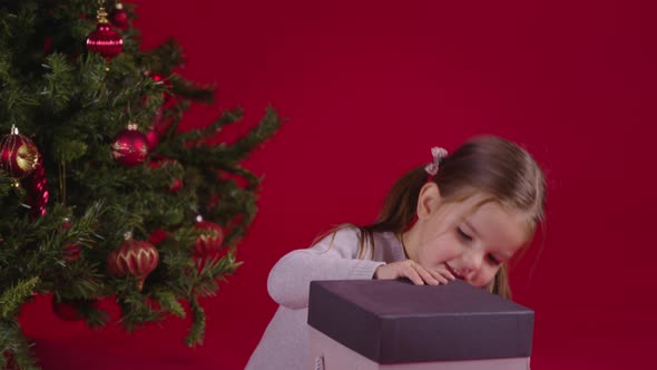 Little Excited Girl Opening Christmas Gift Box Near Xmas Tree on Red Background