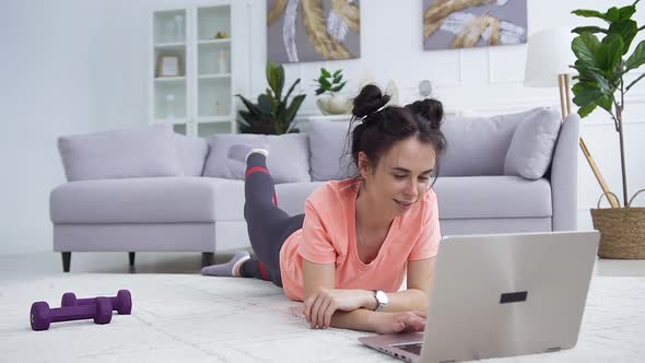 Smiling Young Brunette in Sportswear Lying on the Carpet in Front of the Laptop