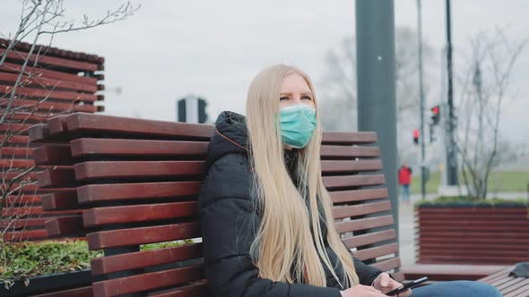 Blonde Girl in a Medical Mask Sitting on the Bench and Using Smartphone