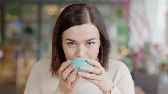 Brunette Caucasian Young Woman with Hazel Eyes Drinking Coffee and Looking at Camera