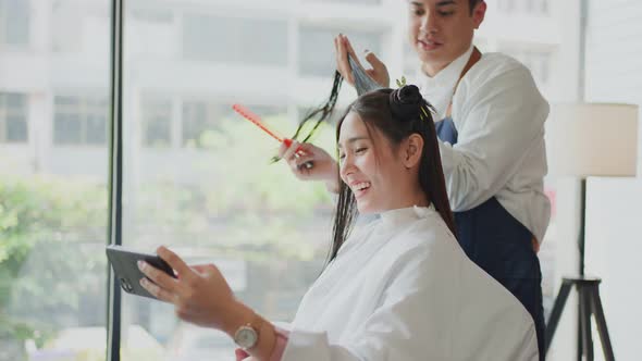 Asian beautiful woman customer watch video on smartphone while getting treatment from hairdresser.