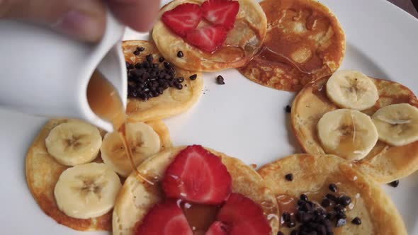 Pouring Maple Syrup in a circular motion over an elegant arrangement of pancakes with sliced strawbe