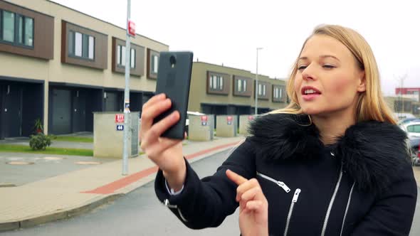 Young Blonde Woman Takes Selfie Photos on Street, Buildings in the Background