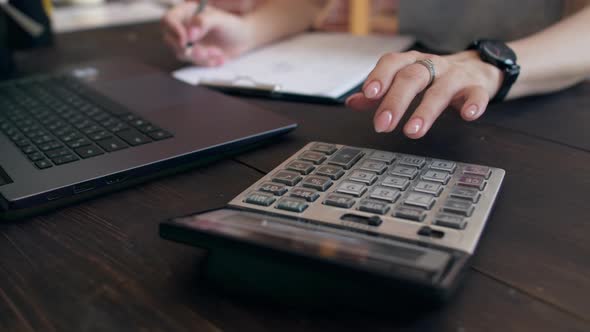 Woman Sits at an Office Table with Paper Tablet and a Laptop and Counts on Calculator Holding a Pen