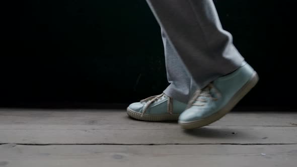 Feet in Blue Sneakers and Gray Pants Walk on Unpainted Boards Against a Black Wall