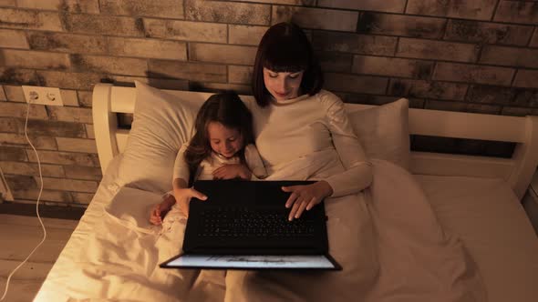 Lovely Mother and Daughter on White Bed Looking at Laptop Together Watching Videos and Having Fun