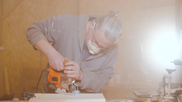 Senior Old Bearded Worker Works with an Electric Jigsaw and Processes Wooden Products