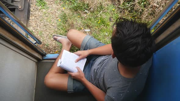 Top View of Young Man Sits Near Entrance of Moving Train and Looks Out at Nature. Male Tourist Rides