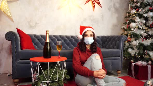 Young Woman in Medical Mask and Santa Claus Hat Sitting on Floor Near Sofa in Room