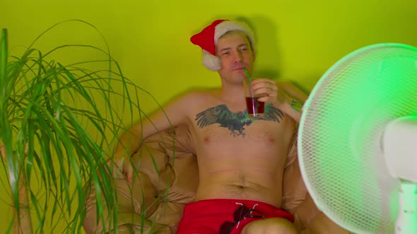 Happy Man in Santa Hat with Bare Torso Drinks Cocktail Through Straw Sitting in Armchair Before Fan