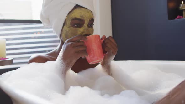 Smiling african american woman with towel and mask taking bath and drinking coffee in bathroom