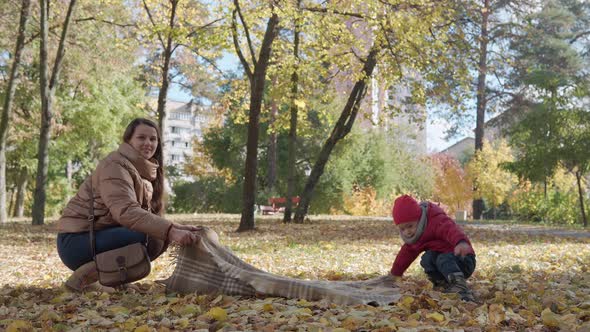 Young Mother Babysitter Picnicking with Little Kid Child Boy Spreads Plaid on Ground Yellow Fallen