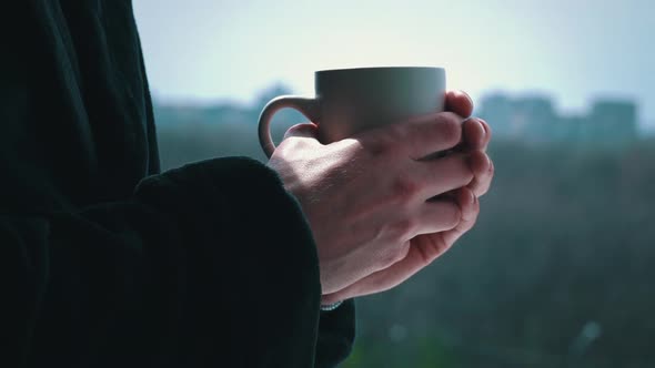 Man Drinks Coffee From Cup in Black Bathrobe at an Open Window in the Morning