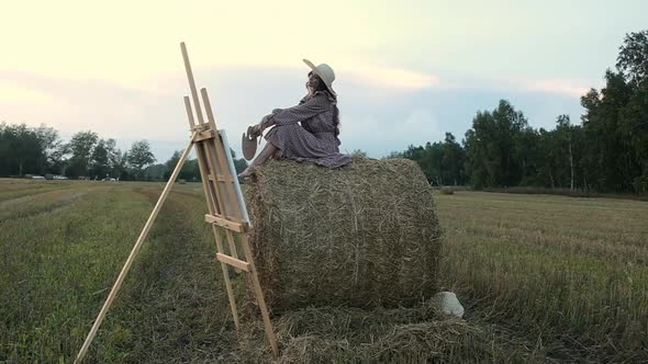 Happy woman in sundress and hat is sitting on haystack looking  painted picture