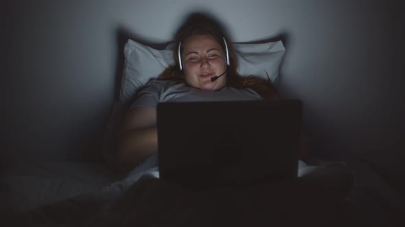 Young Obese Woman Making Video Call with Headset and Laptop in Bed at Night