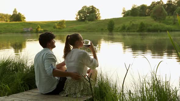 Happy Young Couple Hug Each Other and Drink Red Wine on the Lake Shore