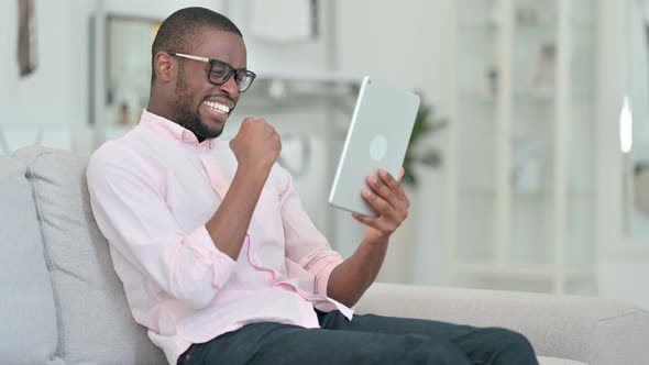 Excited African Man Celebrating Success on Tablet at Home 