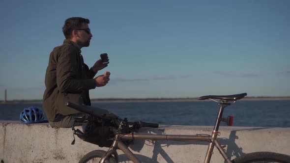 Cyclist Sits on Concrete Fence Near Sea and Drinks From Disposable Cup of Coffee and Eats Sandwich