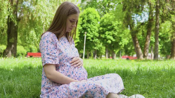 Pregnant Woman in a Summer Dress with a Floral Print Walks in the Park