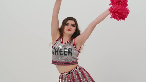 Young Cheerleader with Red Pompoms in Uniform is Dancing on White Background in Studio