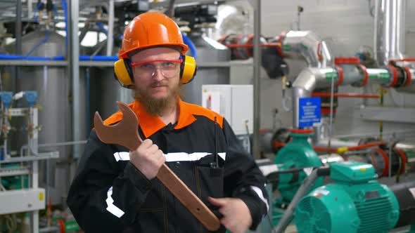 Technological Mechanic in an Orange Helmet Headphones and Goggles Holds a Large Wrench in His Hands