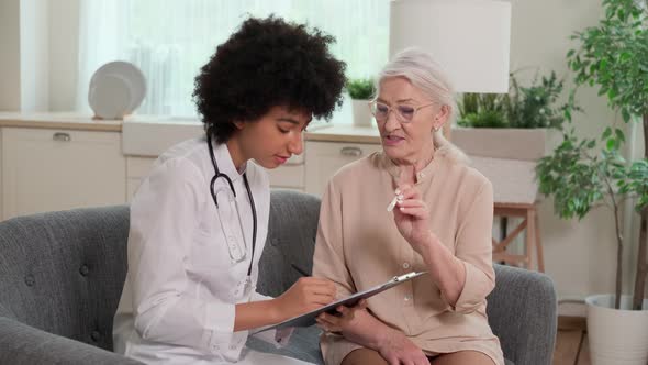 Afro American Woman Doctor Looking at Thermometer Reading at Senor Woman Sitting on Sofa at Home