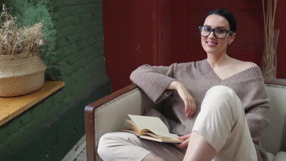 Stylish Young Woman in Glasses with a Book By the Window Sits on the Sofa