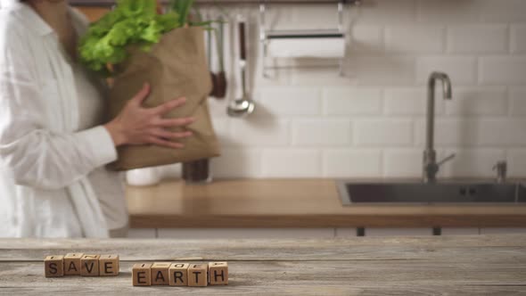 Save The Earth Lined With Cubes On Kitchen Table. Woman Carries Paper Bag, Places It Near