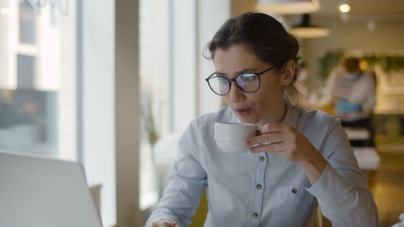 Young Businesswoman in Eyeglasses Working on Laptop Drinking Coffee in Cafe on Lunch Break