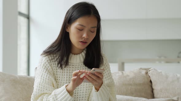 Young Asian Woman Sitting on the Sofa in the Living Room Holding a Smartphone in Her Hand Rejoicing