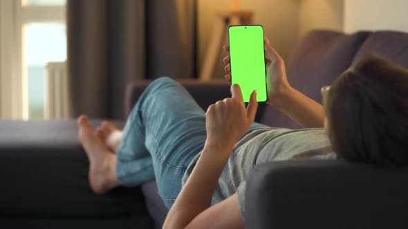 Woman at Home Lying on a Sofa and Using Smartphone with Green Mock-up Screen in Vertical Mode. Girl