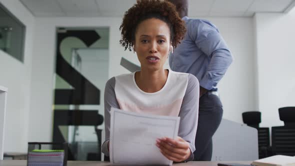 Mixed race businesswoman sitting at desk, holding documents and talking during video call in office