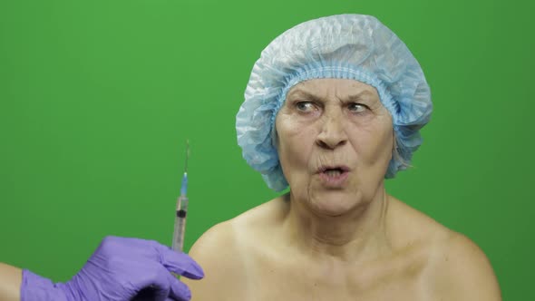 Smiling Elderly Female Lady in Protective Hat Scared of Syringe with Medicines