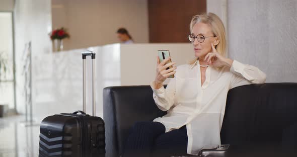 Senior Businesswoman Having Video Call on Smartphone Sitting in Hotel Lobby