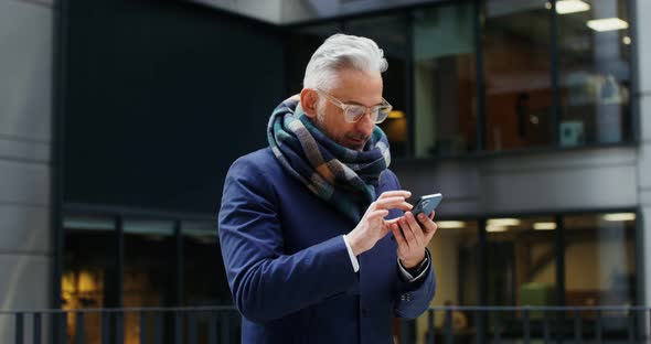 A Grayhaired Man Uses a Mobile Phone Standing in Business Center of the City