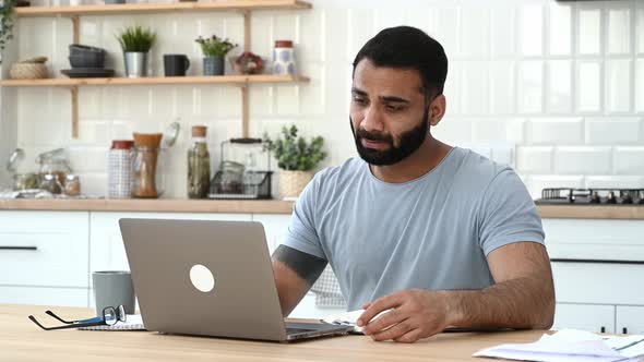 Positive Modern Indian Man Freelancer Designer or Student Sitting at a Kitchen at Home Studying or