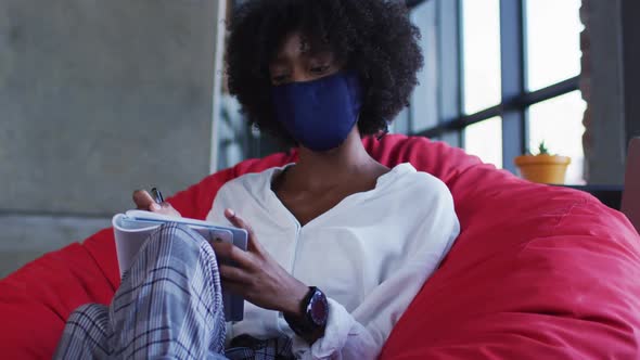 African american woman wearing face mask sitting in cafe writing in notebook