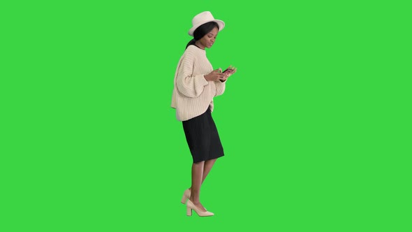 Stylish African American Woman in Knitwear and Hat Using Her Phone While Walking on a Green Screen