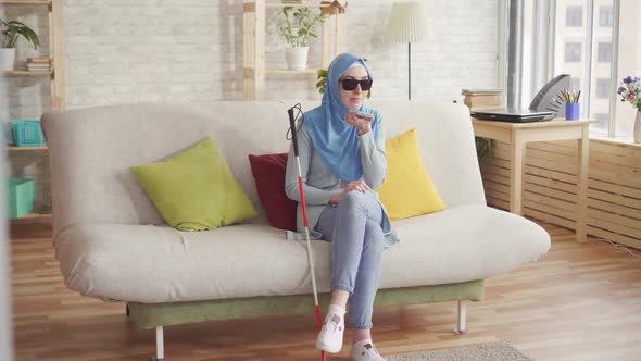 Beautiful Blind Muslim Girl with a Cane in Hijab at Homeenjoys Smartphone Sitting on the Couch