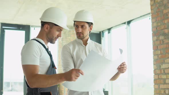 Two Young Engineer in a Safety Helmets Standing at Building Site Holding a Blueprint of the alt