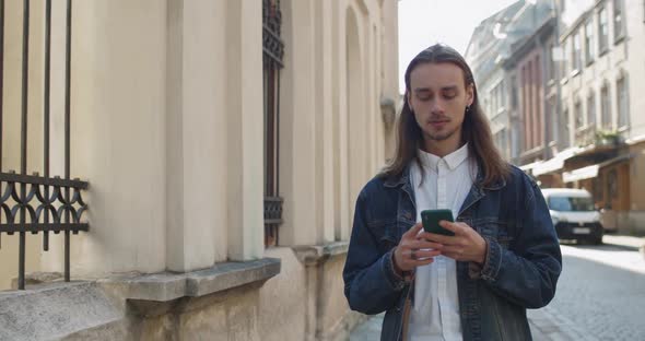 Young Long Haired Guy Using His Smartphone and Looking Forward at Old City Street, Bearded Hipster