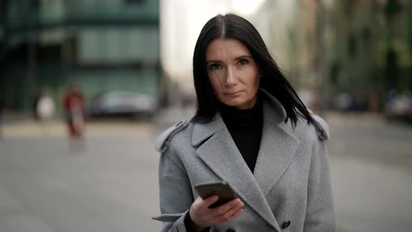 Portrait of a Brunette in a Gray Coat and with a Phone in Hand Against the Background of a Blurry