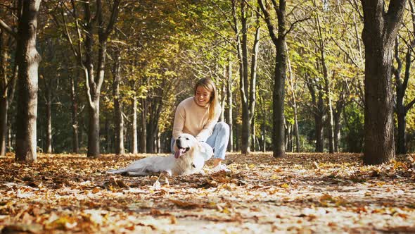 Young Gorgeous Female in Casual Outfit is Sitting on Walkway of Autumn Park Smiling Stroking Her Dog