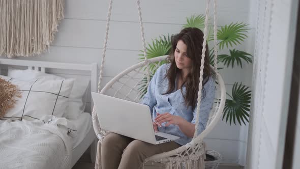 Freelancer Works at Home in a Comfortable Hanging Chair. Girl Working on a Laptop Lying on Her Lap