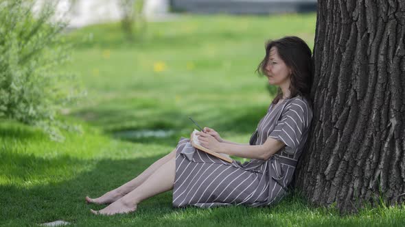 Pretty Adult Woman is Sitting Near Tree with Book Resting and Dreaming