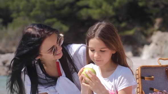 Mom and daughter eat an apple and laugh on Family picnic near sea on sunny summer day.