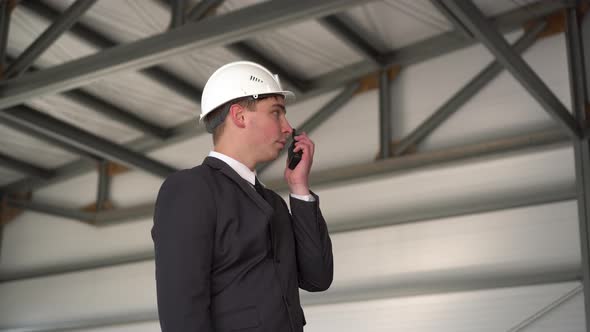 A Young Man in a Helmet Speaks on a Walkie-talkie at a Construction Site. The Boss in the Suit Looks